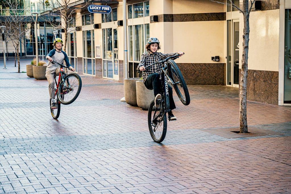 Two teenage boys performing bicycle stunts outdoors in a modern urban setting during the day.